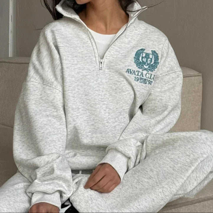 Woman wearing a gray tracksuit with a logo, sitting on a couch.