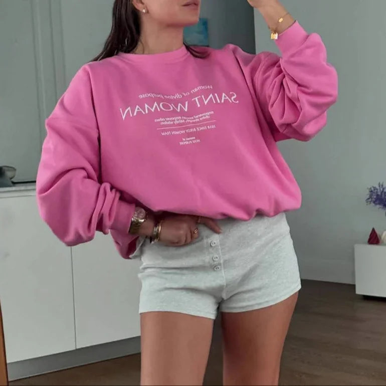Woman taking a mirror selfie wearing a pink sweatshirt with text and gray shorts in a room.