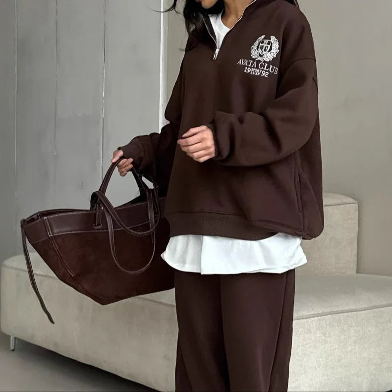 Person wearing a brown tracksuit with a logo, holding a matching bag indoors.