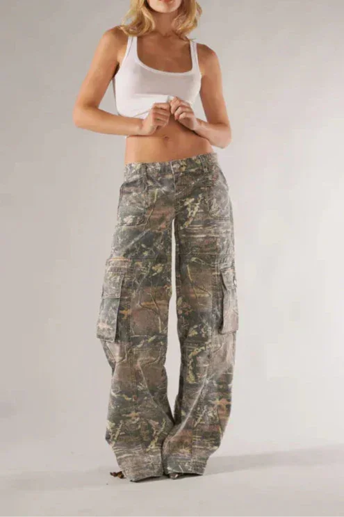 Woman wearing camouflage pants and a white top on a plain background