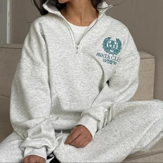 Woman wearing a gray tracksuit with a logo, sitting on a couch.