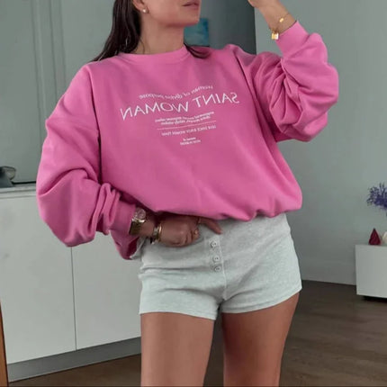 Woman taking a mirror selfie wearing a pink sweatshirt with text and gray shorts in a room.
