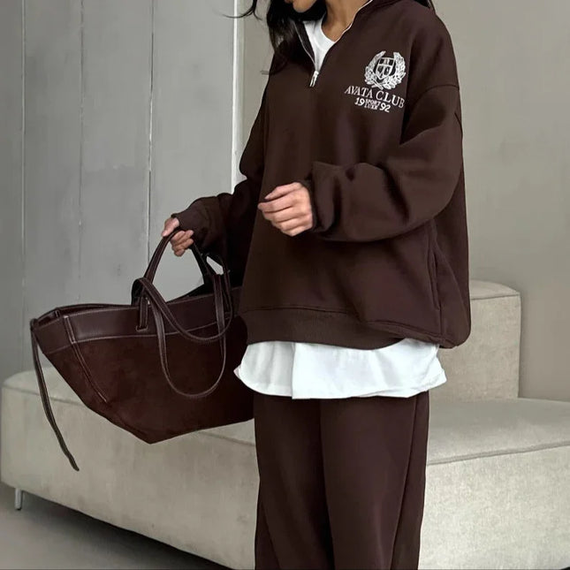 Person wearing a brown tracksuit with a logo, holding a matching bag indoors.