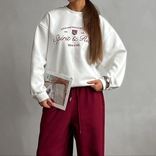 Person wearing a white sweatshirt with text and red pants, holding a magazine against a gray background