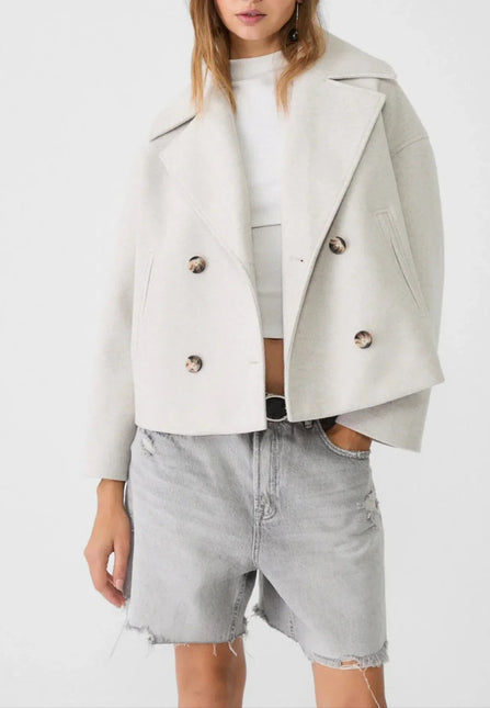 Woman wearing a light gray jacket over a white top and denim shorts on a white background
