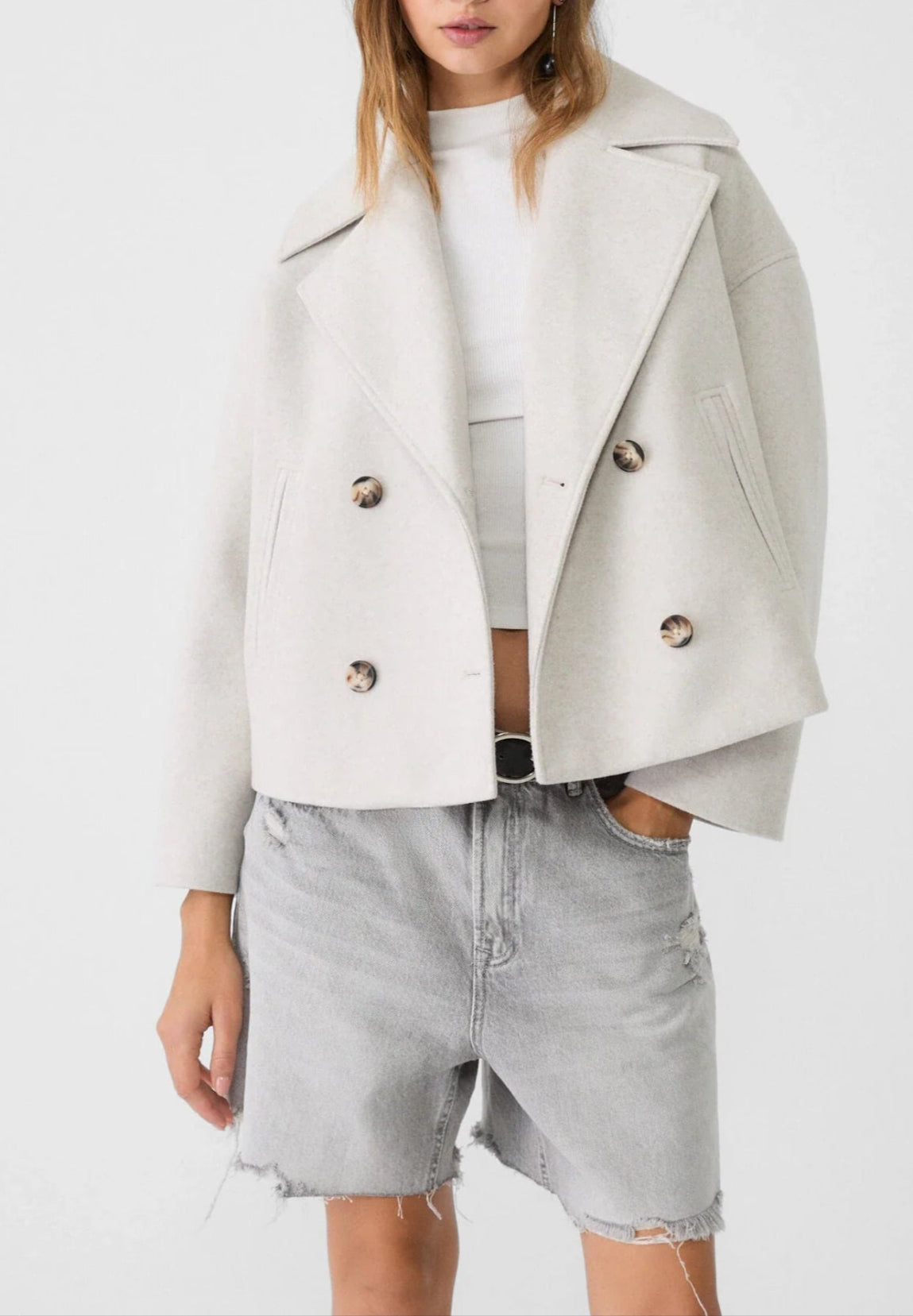 Woman wearing a light gray jacket over a white top and denim shorts on a white background